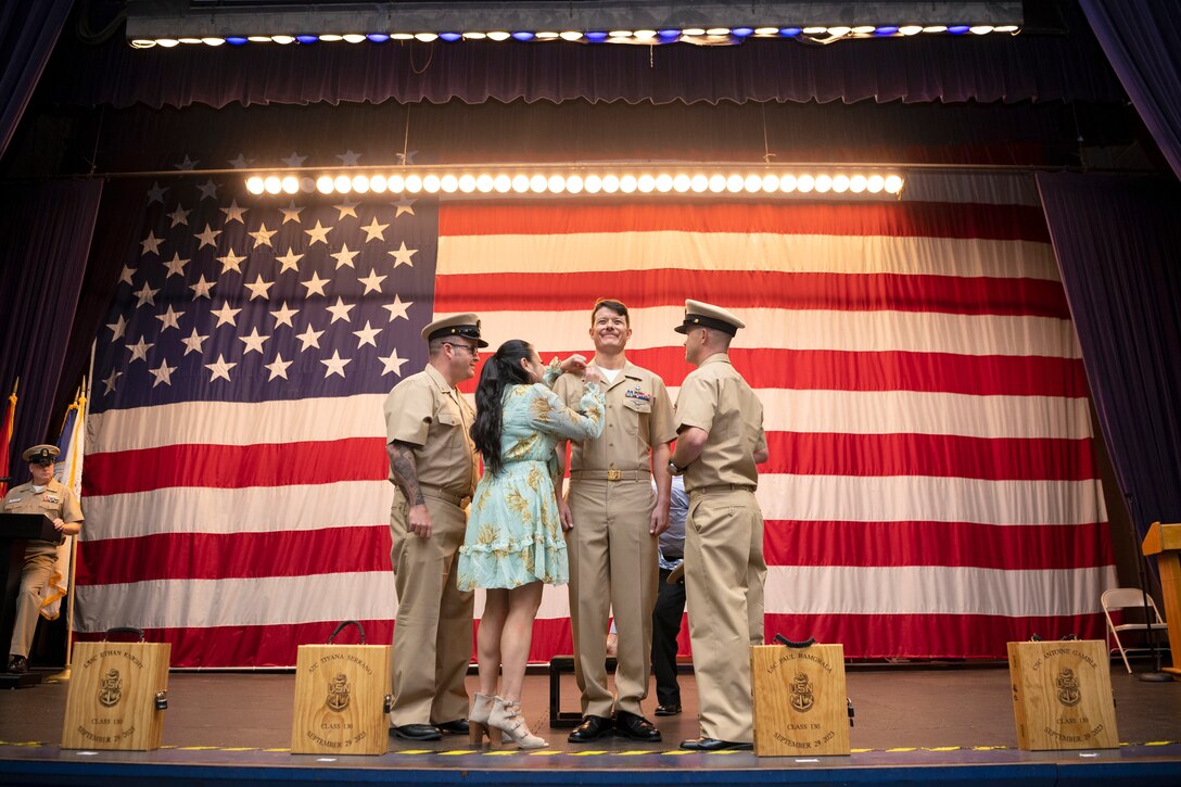 A loved one puts a pin on a sailor’s collar as two fellow sailors stand nearby as spotlights shine on an American flag in the background.