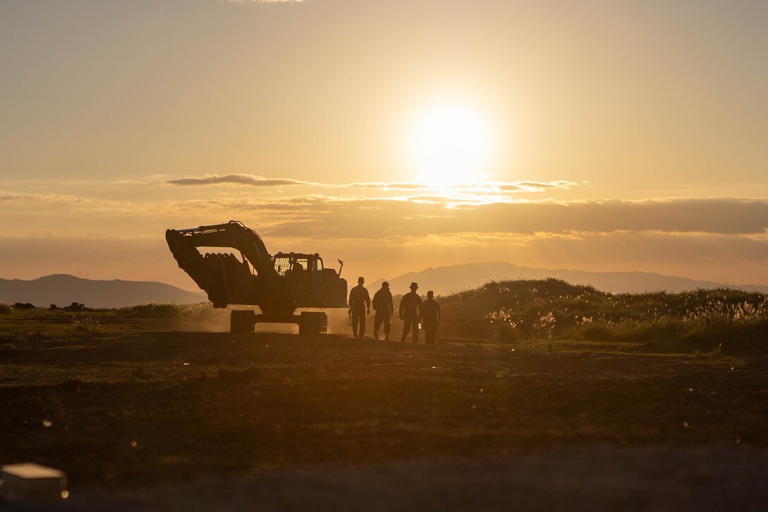 Marines stand near a forklift at a construction site. They are illuminated by sunlight.