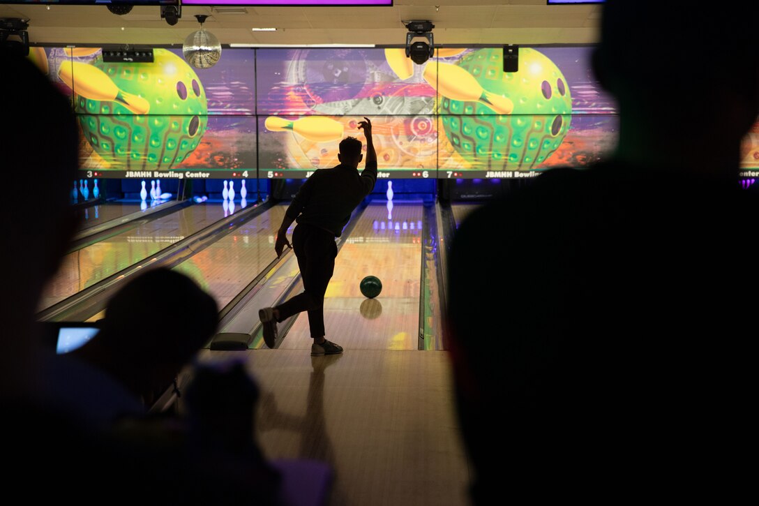 A soldier photographed in silhouette releases a bowling ball down a lane as others watch. There is one pin left to hit.