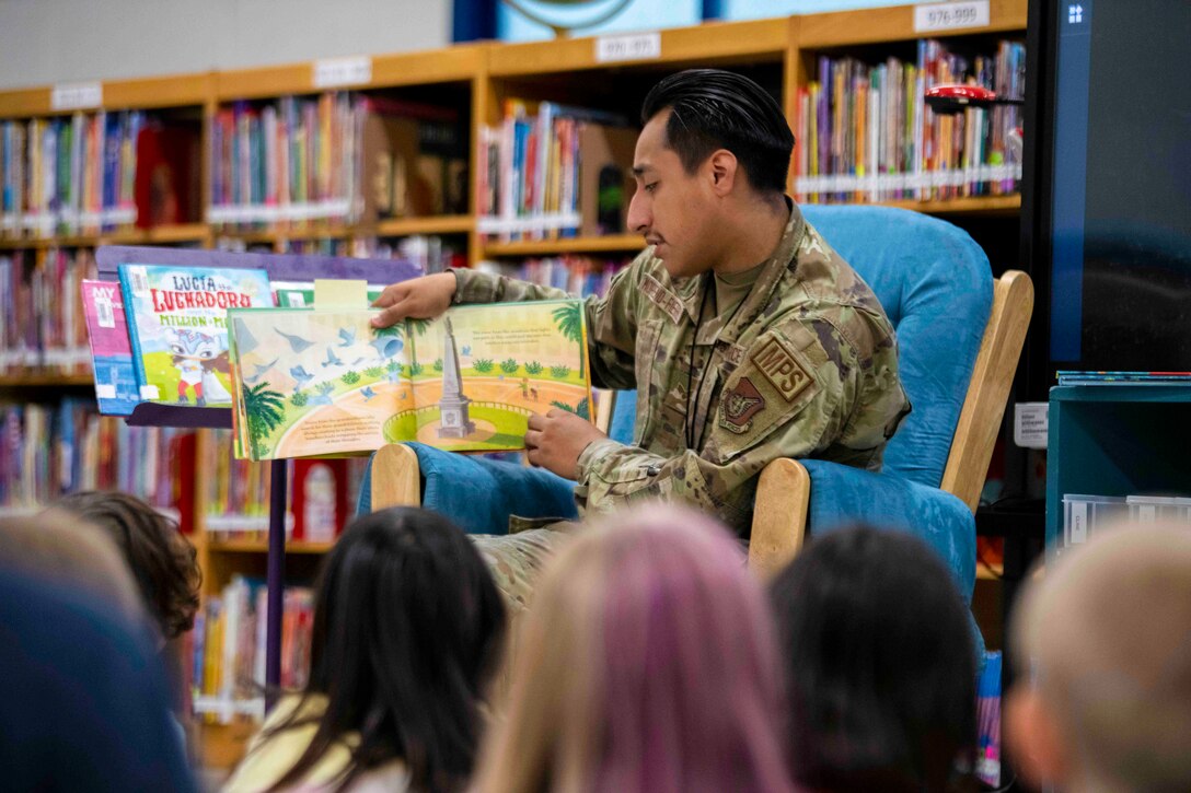 An airman sits in front of a group of children while reading a book.