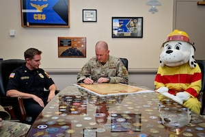 U.S. Air Force Col. Joshua DeMotts, 99th Air Base Wing commander, sits at a desk as he signs the 2023 Fire Prevention Week Proclamation with members of the 99th Civil Engineering Squadron.