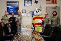 From left, U.S. Air Force Senior Master Sgt. Jeremiah Gates, 99th Civil Engineering Squadron (CES) deputy fire chief, Mr. Aaron Grindland, 99th CES assistant fire chief, U.S. Air Force Col. Joshua DeMotts, 99th Air Base Wing (ABW) commander, Sparky, U.S. Air Force Lt. Col. Jenny Gibson, 99th CES commander, and U.S. Air Force Chief Master Sgt. Adrienne Warren, 99th ABW command chief, stand for a photo while holding to display the 2023 Fire Prevention Week Proclamation.