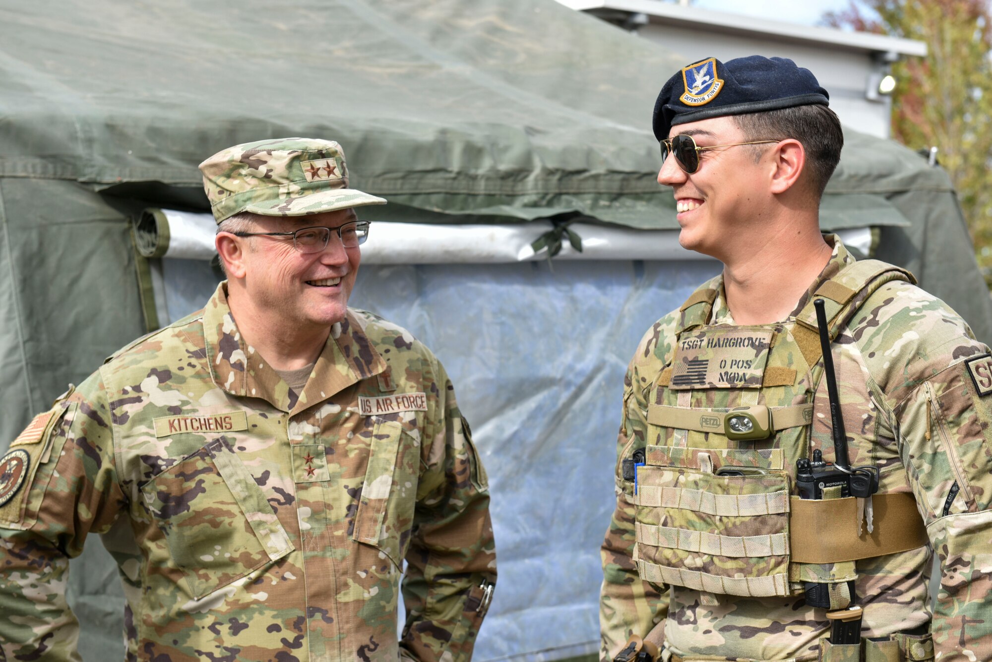 Maj. Gen. Randall Kitchens stands next to Tech Sgt. Ashton Hargrove outside near the main gate. They are both smiling at a joke that was made in their conversation.