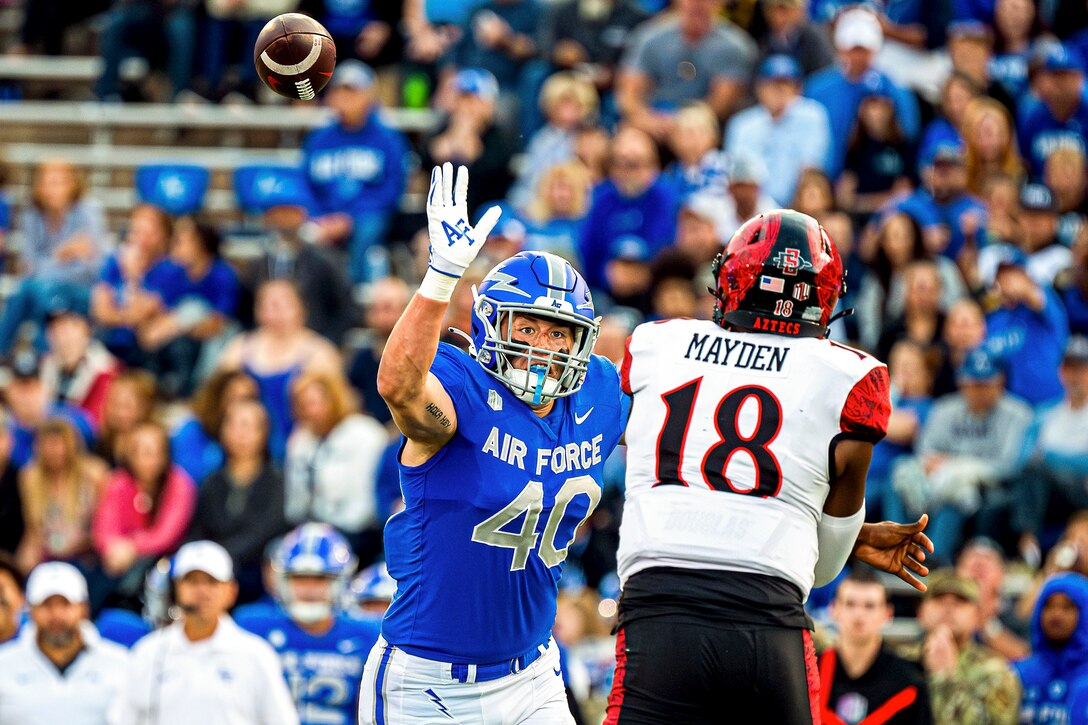 An Air Force football player raises their hands to block a ball thrown by an opponent as fan watch from the stands in a blurred background.