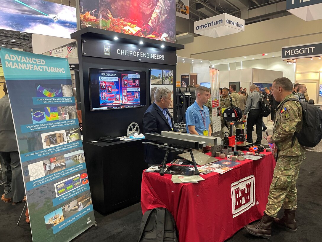 Two people stand behind red tabled booth in a crowded convention center speaking with a person in green, brown and tan military uniform.