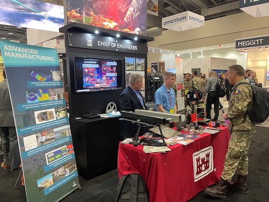 Two people stand behind red tabled booth in a crowded convention center speaking with a person in green, brown and tan military uniform.