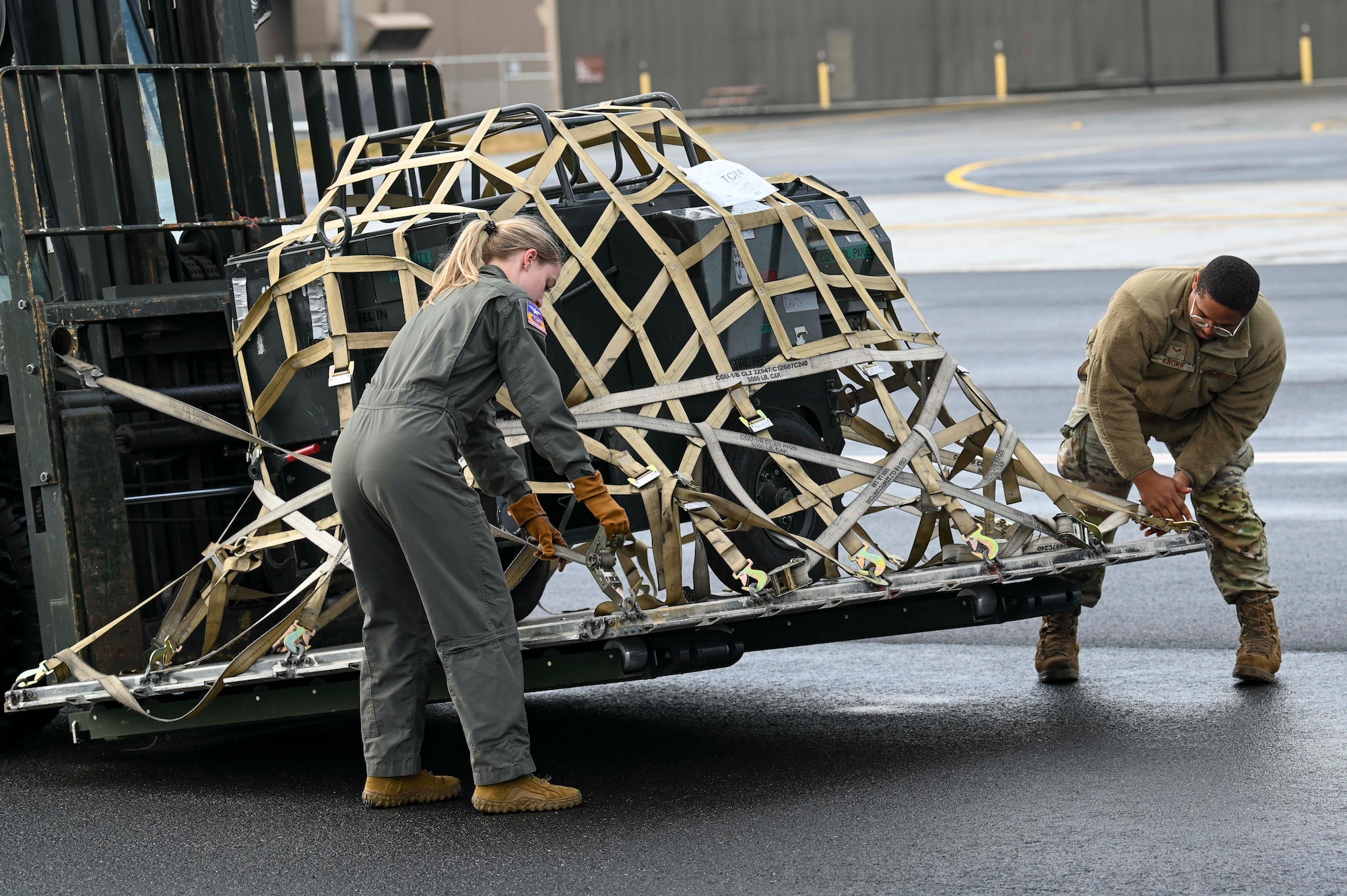 Two Airmen adjust a net that is on top of cargo holding it in place on the pallet of the forklift