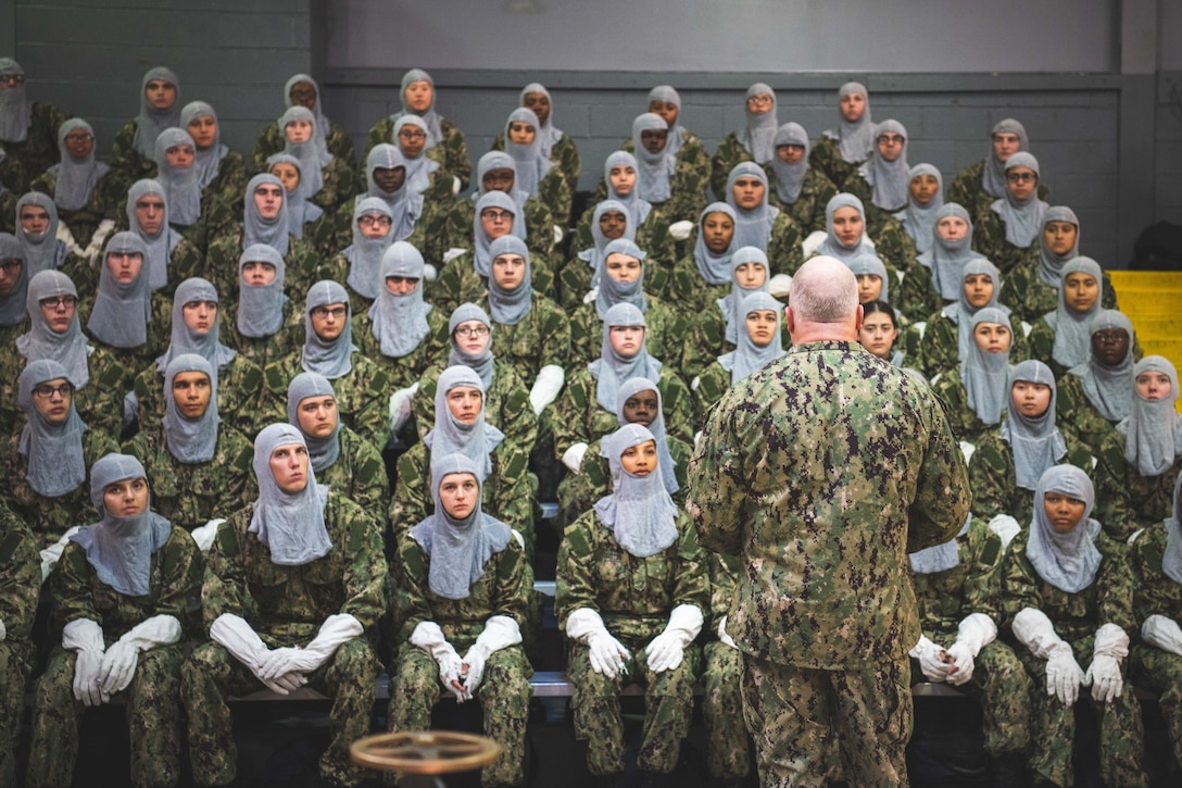 Sailors wearing white hoods and gloves sit and listen to a lecture.