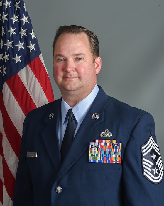 A man in a blue dress uniform stands for a photo in front of an American flag and grey background.