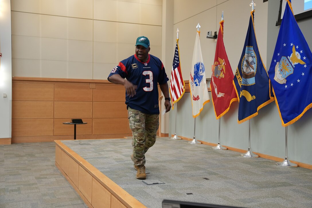 man in Army uniform and football jersey walks on auditorium stage
