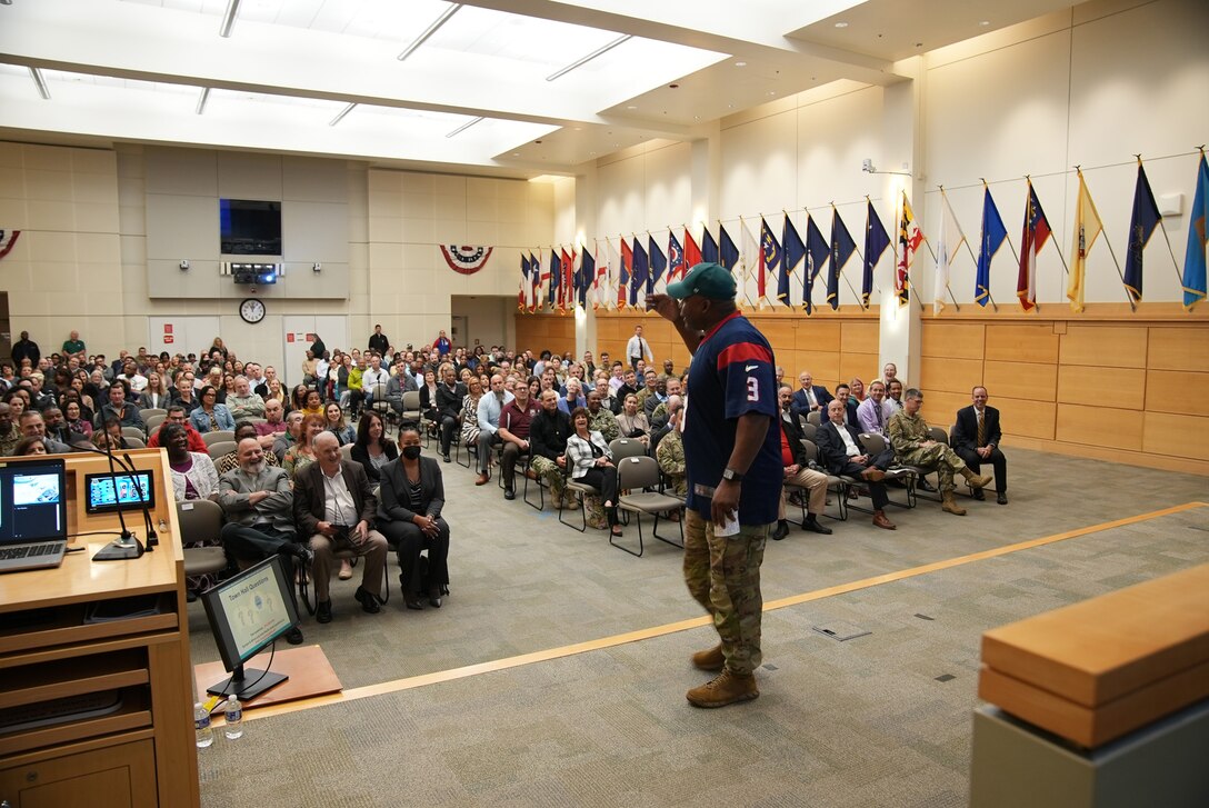 man in Army uniform and football jersey talks to crowd of seated people