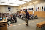 man in Army uniform and football jersey talks to crowd of seated people