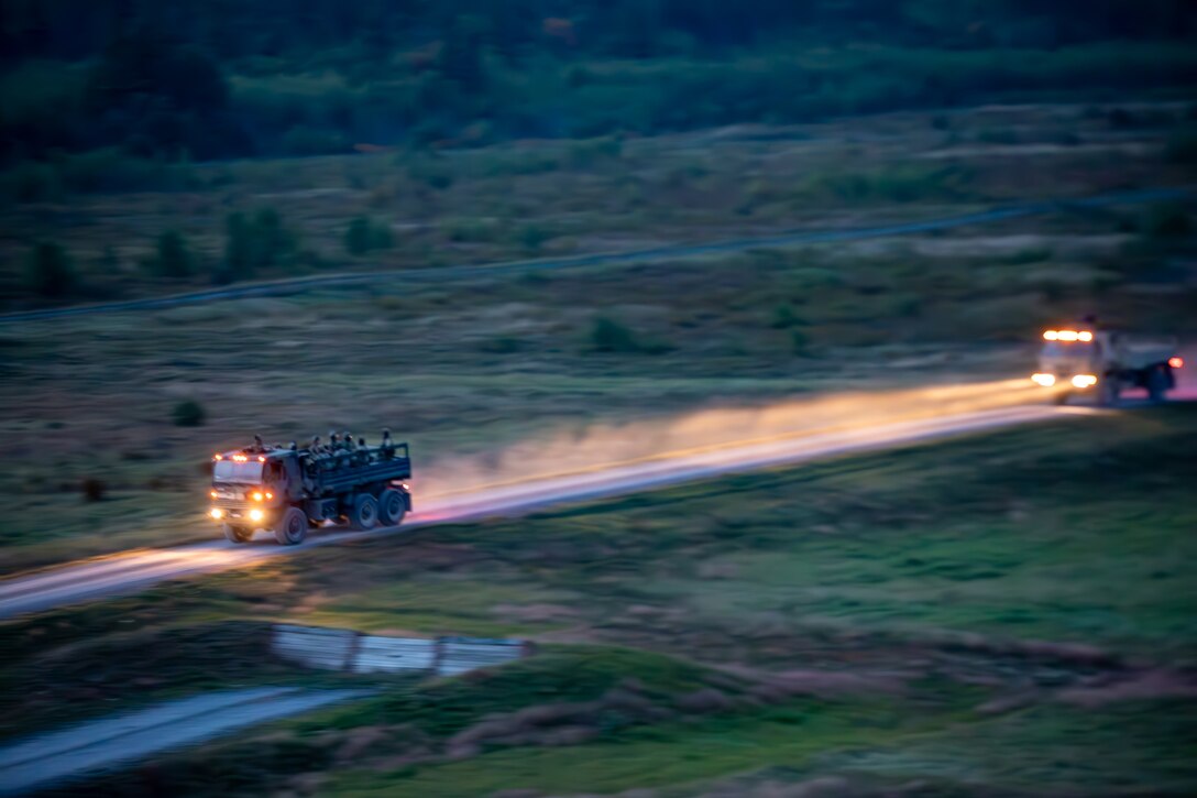 Two military vehicles drive down a road at dusk. The vehicles are surrounded by grassy areas.