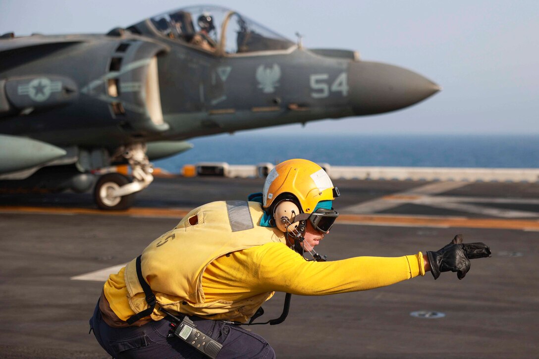 A sailor signals in front of an aircraft on the deck of a ship.