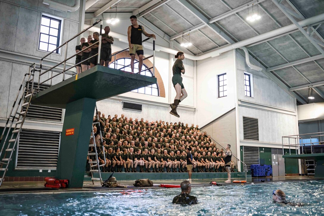 A recruit jumps into a pool in front of recruits sitting in stands and others standing on a diving board above.