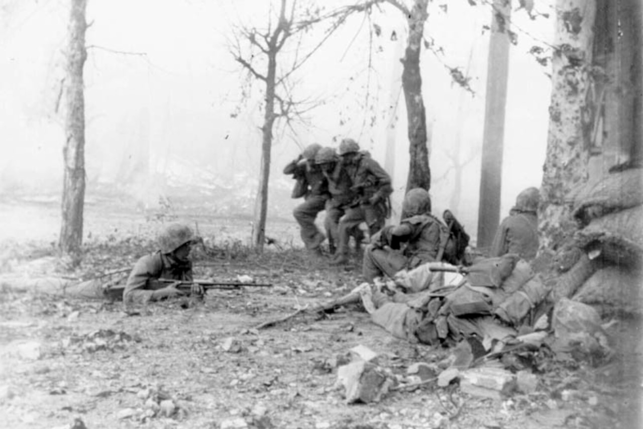 Two soldiers carry another man over their shoulders as others crouch low during battle.