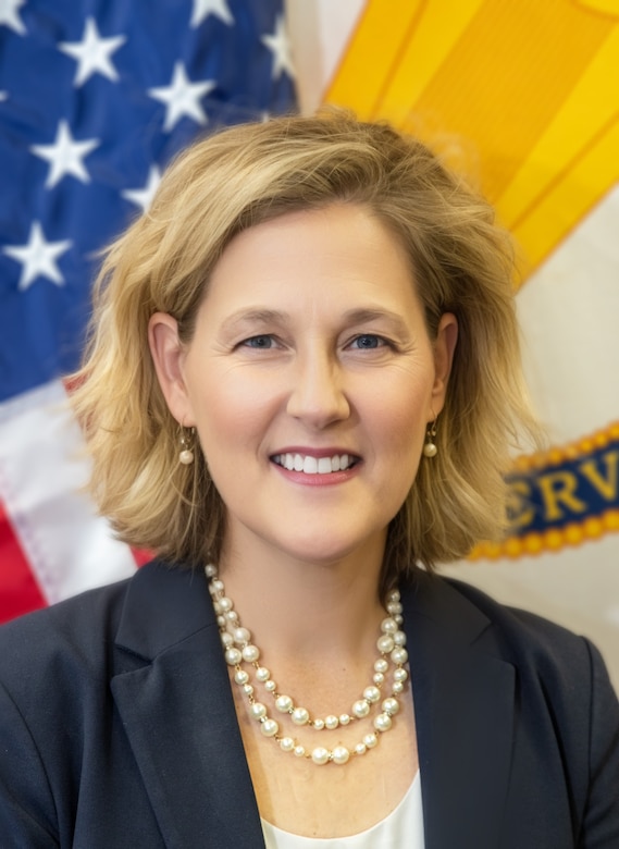 A person, Ms. Tambour L. Eller, with sandy blonde hair, wearing a blue jacket, white pearls and a white top, smiles in front of the U.S. Flag and the flag of the U.S. Army