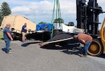 Men assist large propeller to the ground as a forklift crane lowers it to rest on wood beams.