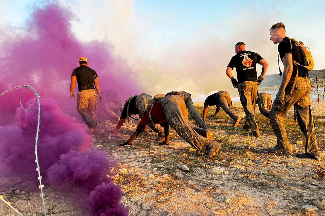 Soldiers move one knee to their chest while holding their hands on rocky soil surrounded by clouds of purple smoke as fellow soldiers watch.