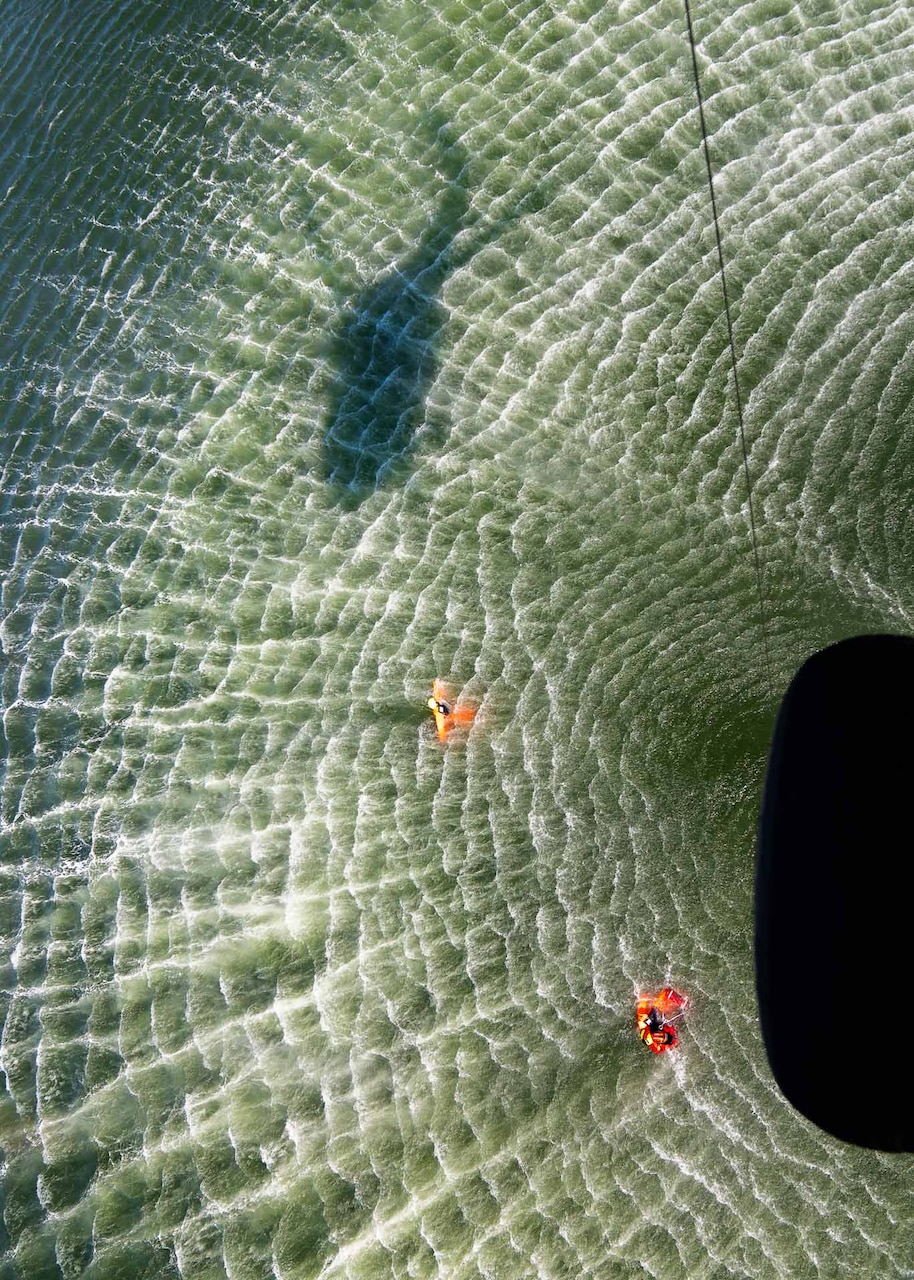 A sailor moves through a body of water as a shadow of a helicopter shines on the water.