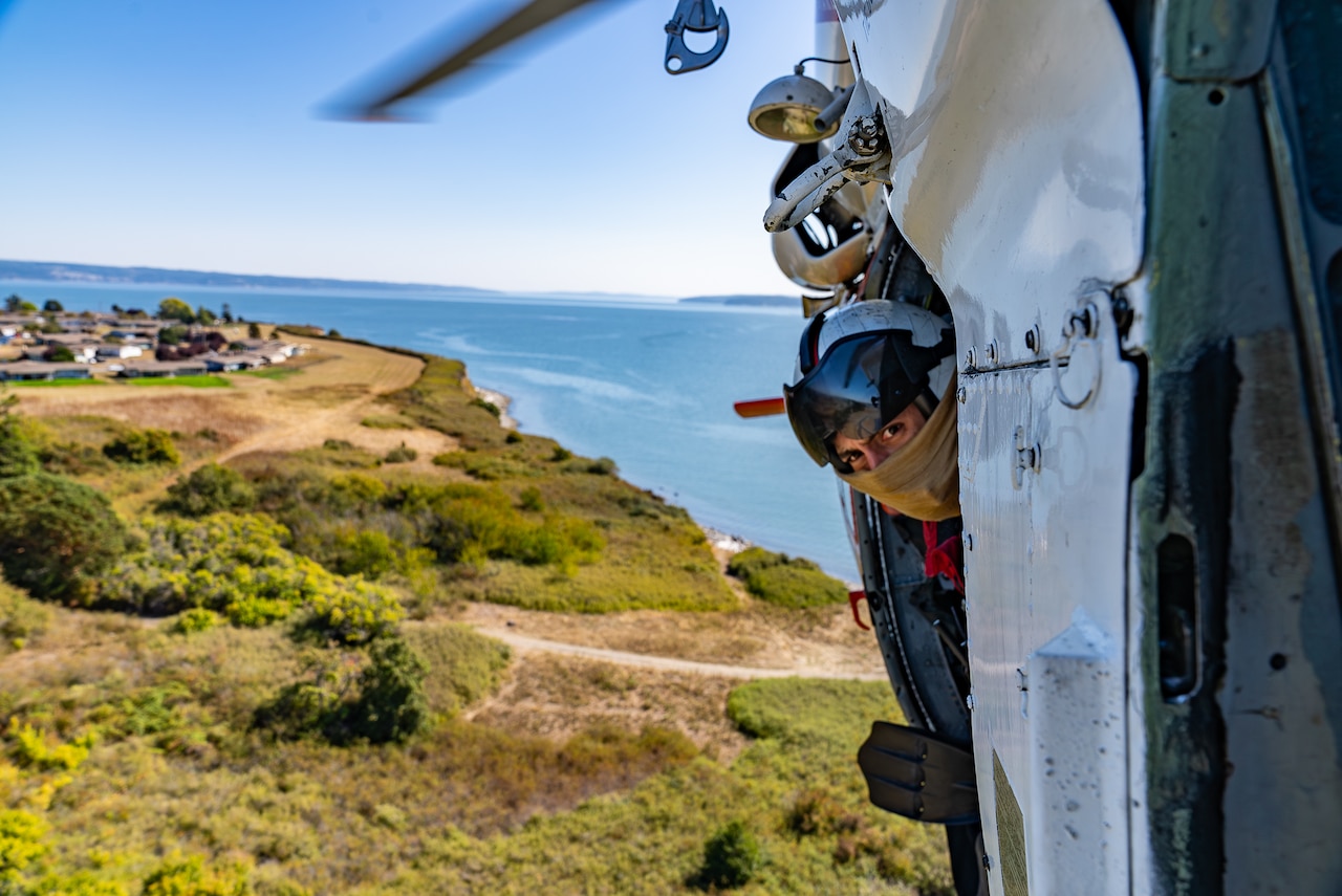 A sailor looks out the door of an airborne aircraft.