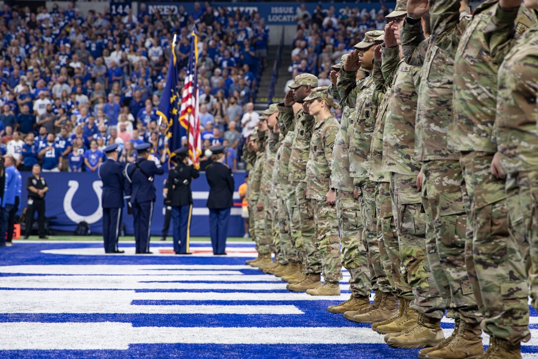 Service members stand in a line and salute in a football stadium.