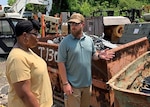 A woman in a tan shirt listens to a man with a beard and green shirt as he talks. he is using one hand to indicate a container of scrap metal.