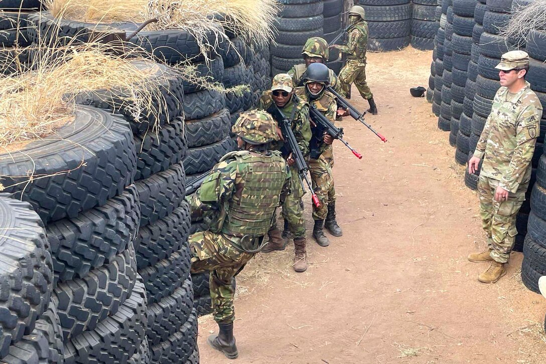 A soldier stands behind foreign troops moving through a maze of tires.