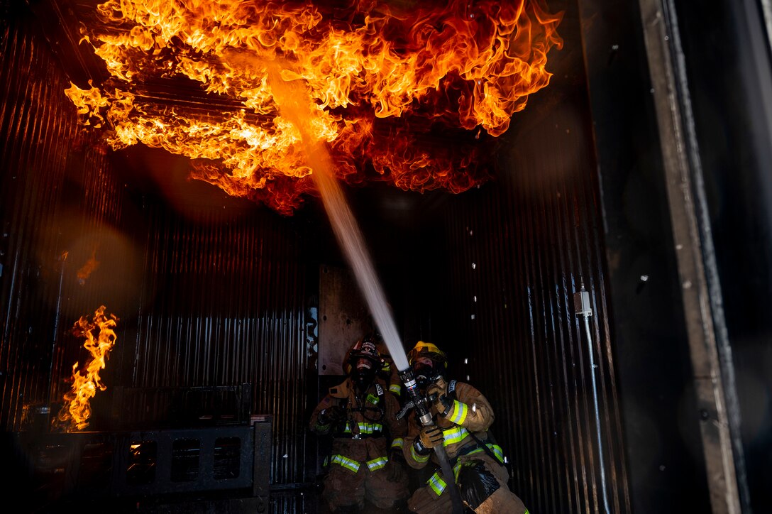 Two Air Force fire protection specialists fight a fire overhead during training. One holds a water hose and sprays it toward the ceiling.
