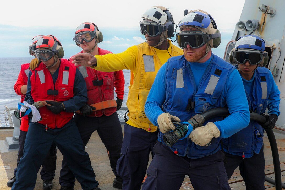 Two groups of sailors wearing red and blue hold onto water hoses as another sailor wearing yellow stands between them pointing ahead.