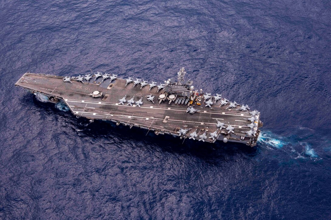 An overhead view of aircraft parked on the deck of a ship as it travels through a body of water.