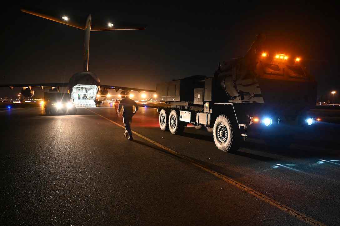 A service member walks toward a military aircraft on a runway at night as a Humvee waits nearby.