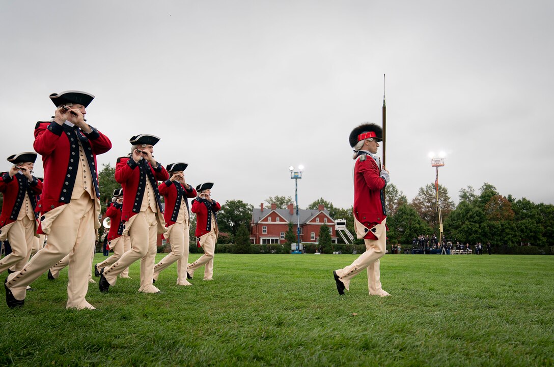 Soldiers in ceremonial uniforms play instruments while marching across a field during a ceremony as the soldier in front holds a sword.