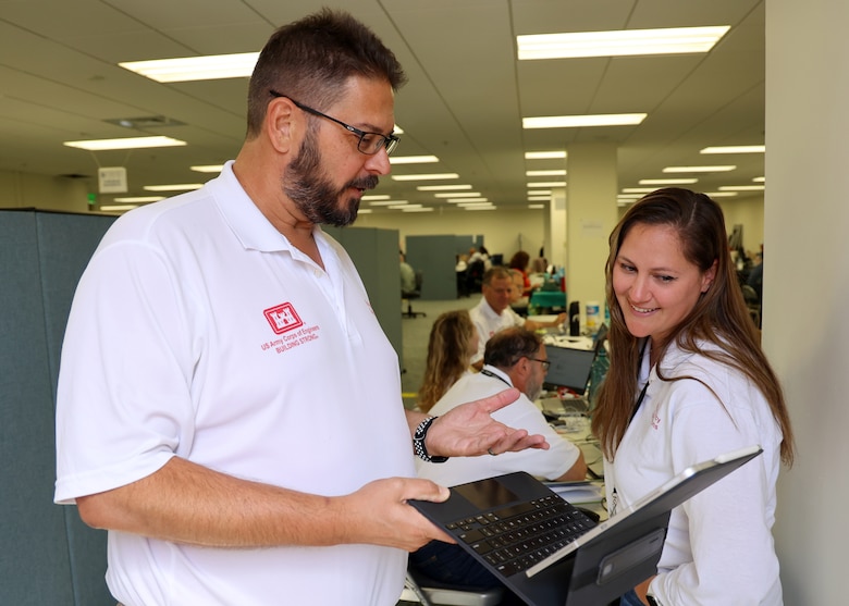 A man and a woman talk in an office setting. The man is holding a tablet computer that the woman is viewing.