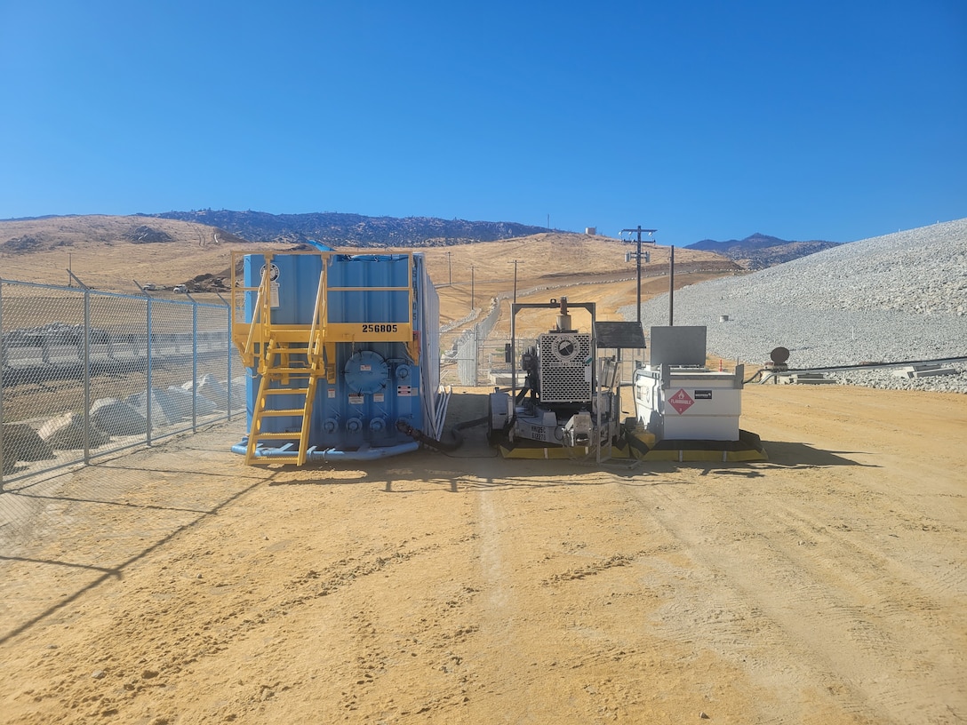 a pumping system at the base of a dam, with a fence to the side and mountains in the background