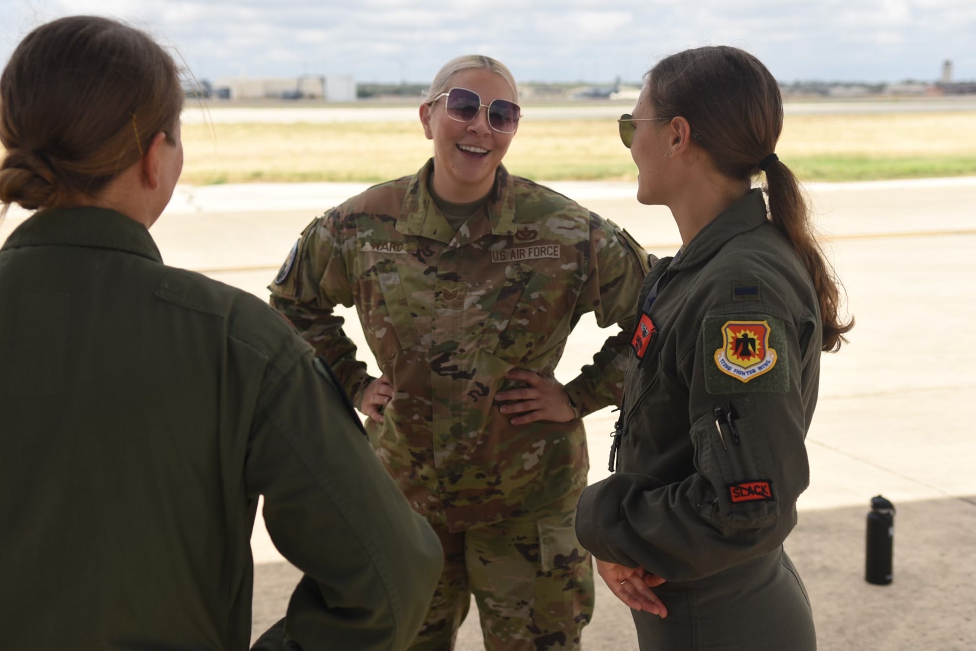 Two women in flights suits and another in OCPs engage in conversation outside.