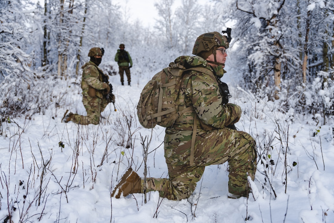 Soldiers kneel in the snow in a wooded area.