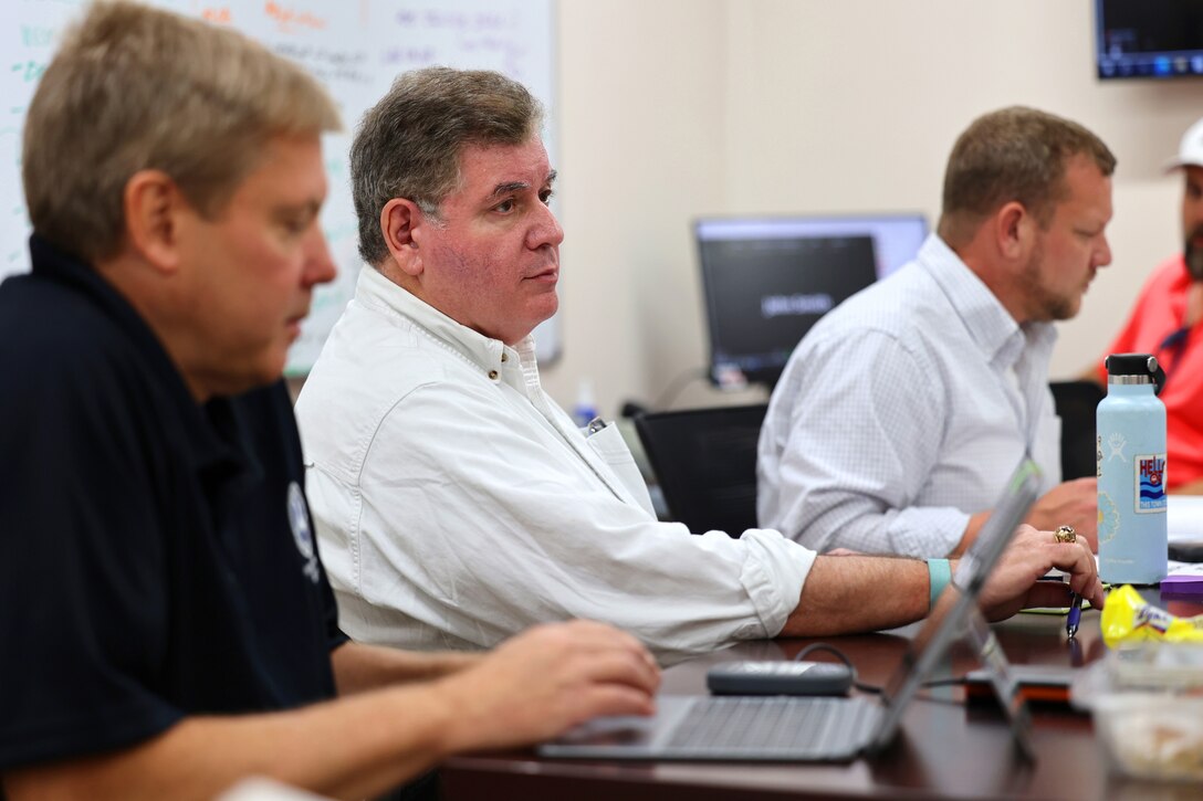 Three men sit in an office setting listening to presenters in a meeting.