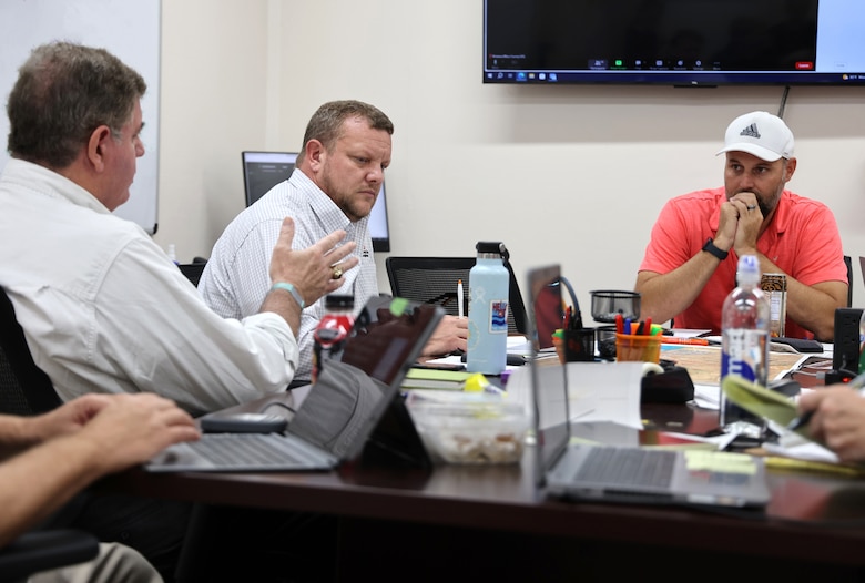 A man speaks to two other men in an office setting.
