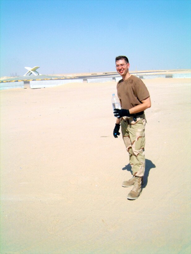 A man in a military uniform stands in the desert with a blue sky in the background.