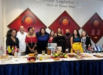 Nine committee members stand behind tables featuring food and displays highlighting Hispanic Heritage Month
