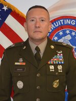 Man in an Army uniform, standing in front of an American Flag and the U.S. Army Recruiting emblem