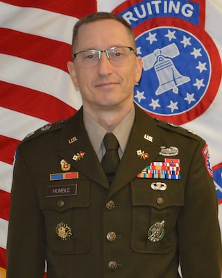 Man in Army uniform, standing in front of the American Flag and the Army Recruiting emblem