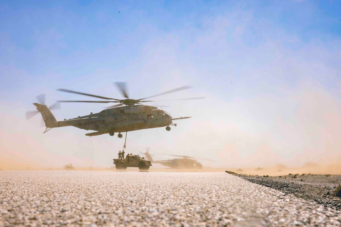 An aircraft hovers above Marines standing on top of a military vehicle; another aircraft is parked behind.