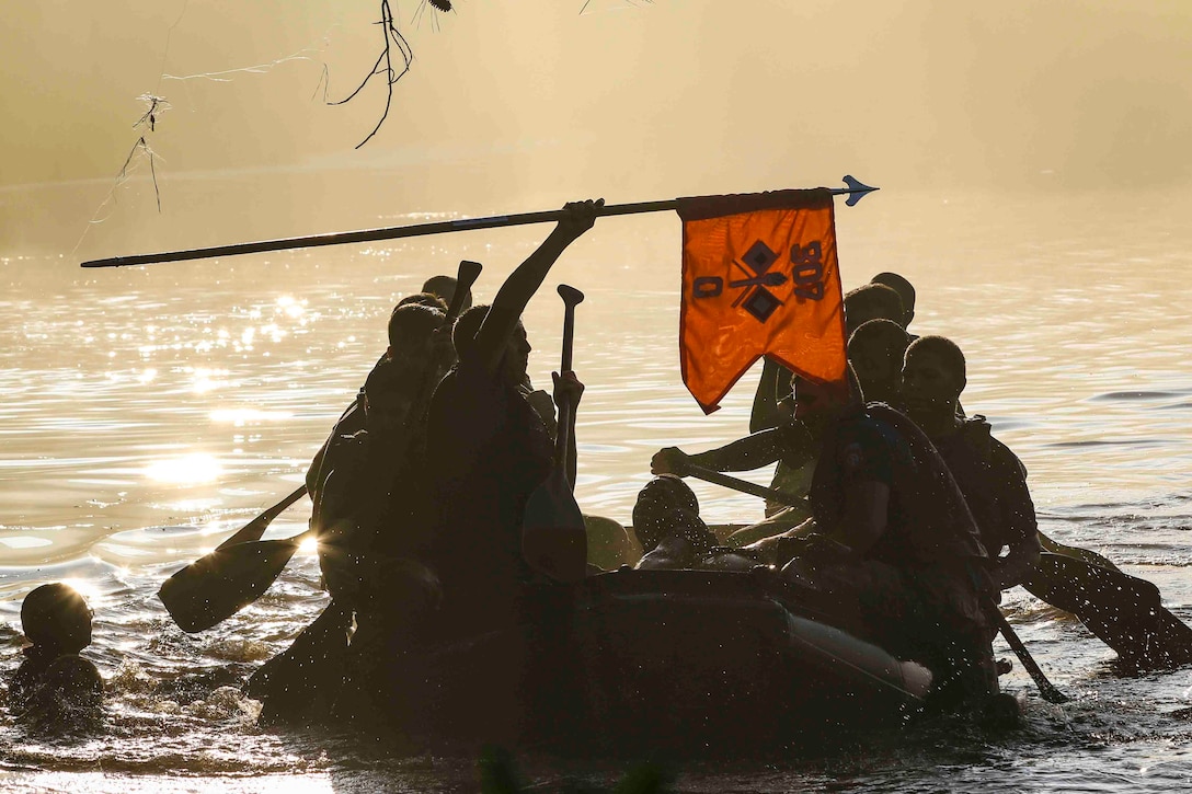 Soldiers shown in silhouette paddle in an inflatable boat across a body of water as one holds a red flag above their heads.