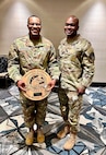 Two men in Army uniforms pose for a picture. The man on the left is holding a wooden plaque.