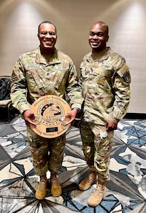 Two men in Army uniforms pose for a picture. The man on the left is holding a wooden plaque.