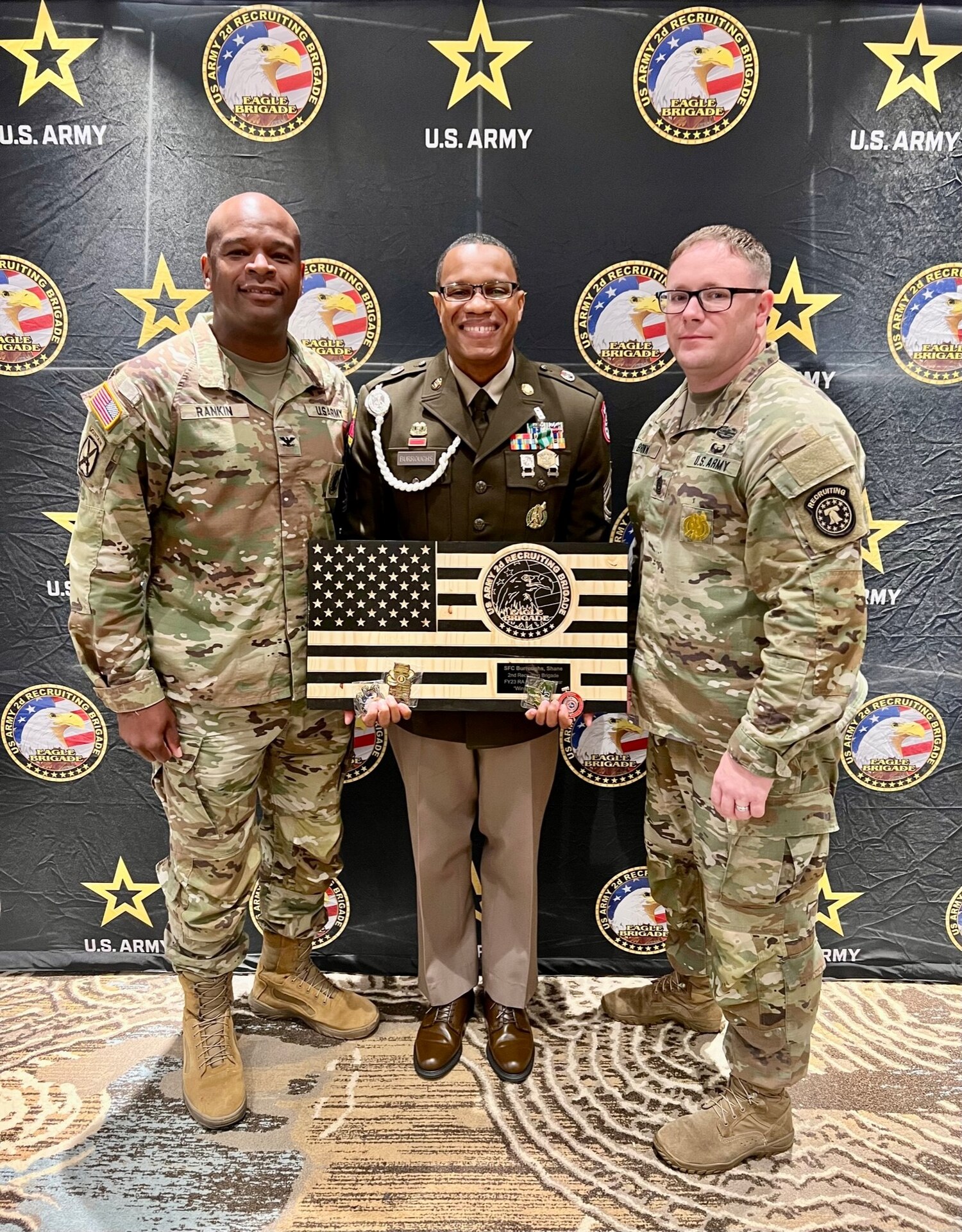 Three men in Army uniforms stand in front of an Army Recruiting branded background. Man in the middle is holding a wooden American Flag