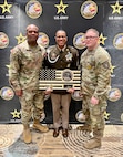 Three men in Army uniforms stand in front of an Army Recruiting branded background. Man in the middle is holding a wooden American Flag