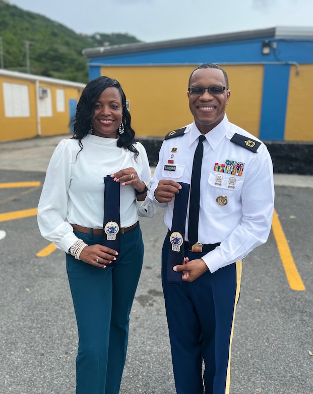 Man on the right in an Army uniform, holding a customized tie and a woman on the left in a white blouse and teal pants holding a similar customized tie